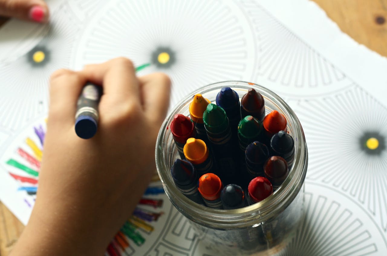 A glass jar with coloured crayons next to a child colouring in patterns on a piece of paper