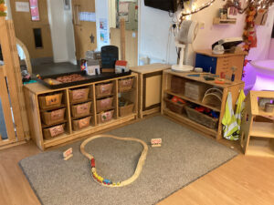 A room with a small wooden train set in the middle on top of a carpet with wooden shelves and baskets next to it
