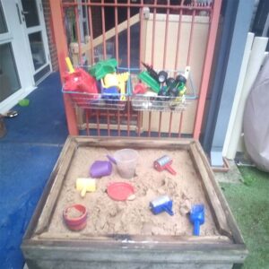 An outdoor sandpit with toys in baskets above attached to a red railing