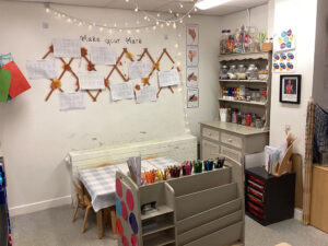 A nursery room with signs and artwork on the wall and a small table and chairs next to some wooden shelves with drawing stuff on